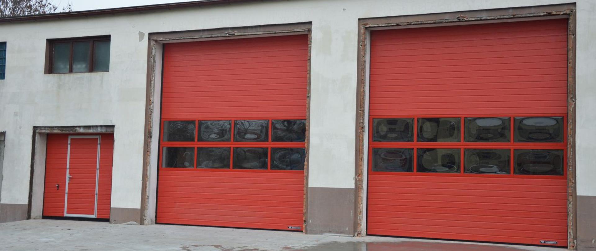 Budynek garażowo-warsztatowy, widok na nowe bramy podnoszone. Bramy koloru czerwonego, dwie z nich (większe) posiadają dwupoziomowe przeszklenie, trzecia - mniejsza (bez przeszklenia) wyposażona jest w drzwi.