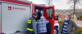 Kolorowe zdjęcie przedstawia na pierwszym planie, czerwony, oznakowany samochód lekki Ochotniczej Straży Pożarnej. Przy samochodzie, na środku widać strażaka Ochotniczej Straży Pożarnej z napisem na plecach Straż, który pomaga starszej osobie pośrodku, wejść do samochodu. Obok stoi inna osoba cywilna. Na zdjęciu widoczne w tle drzewa i zabudowania.