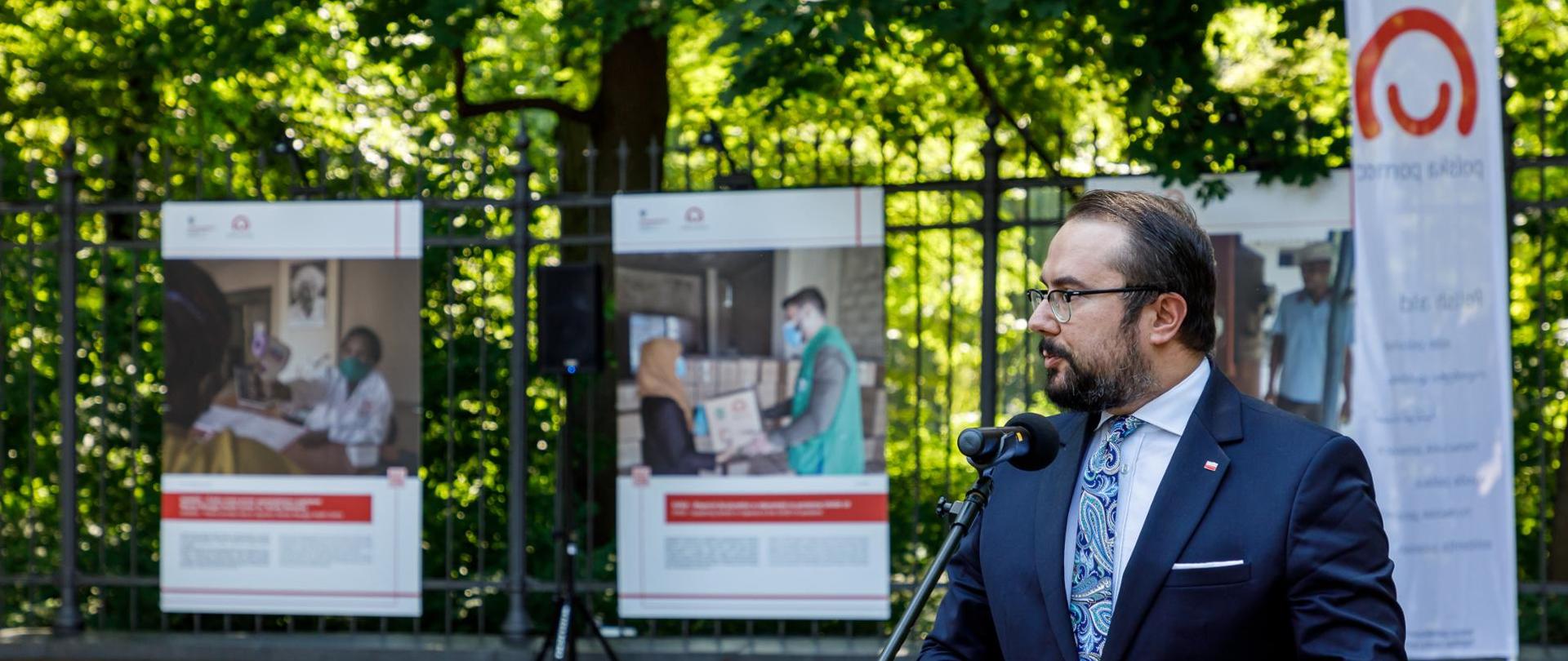 Minister Paweł Jabłoński otwiera wystawę - mężczyzna mówi do mikrofony, w tle na przęsłach ogrodzenia przymocowane są plansze wystawy zawierające zdjęcia oraz tekst