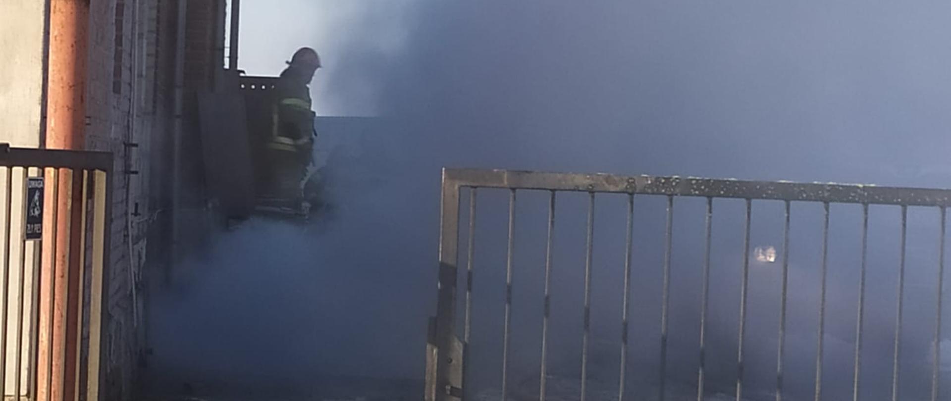 Zdjęcie przedstawia strażaków gaszących pożar w kotłowni i w piwnicy budynku.
W tle budynki.
