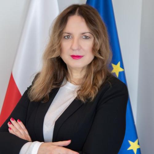 Kobieta stoi z założonymi rękami, ubrana w czarną marynarkę i jasną bluzkę. Ma falowane, jasnobrązowe włosy do ramion i wyraźny makijaż z czerwonymi ustami. W tle znajdują się dwie flagi: po lewej biało-czerwona, a po prawej niebieska z żółtymi gwiazdami.