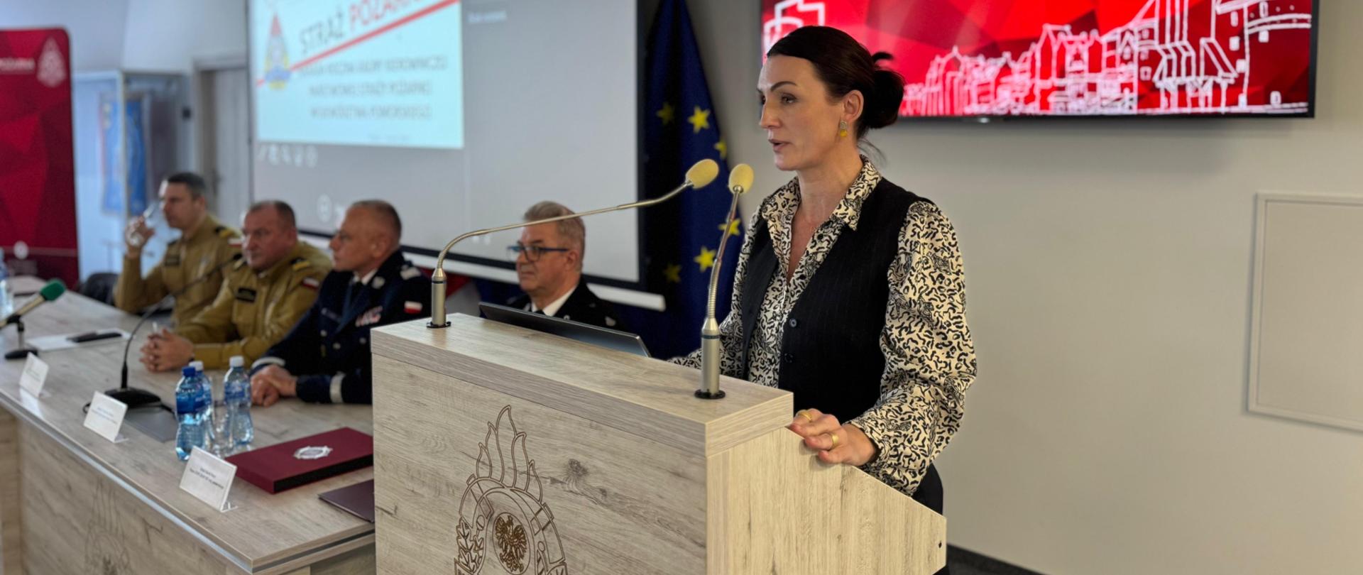 kobieta stoi za pulpitem i przemawia. W tle siedza mężczyźni w strażackich mundurach