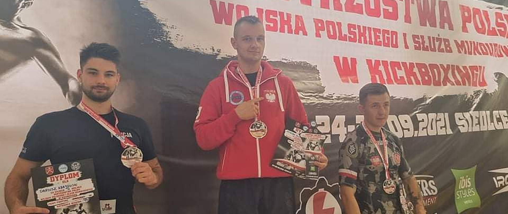 Trzech zawodników stoi na podium na tle baneru z napisem Mistrzostwa Polski Wojska Polskiego i Służb Mundurowych. Na najwyższym stopniu stoi strażak z Elbląga, na szyi ma złoty medal.