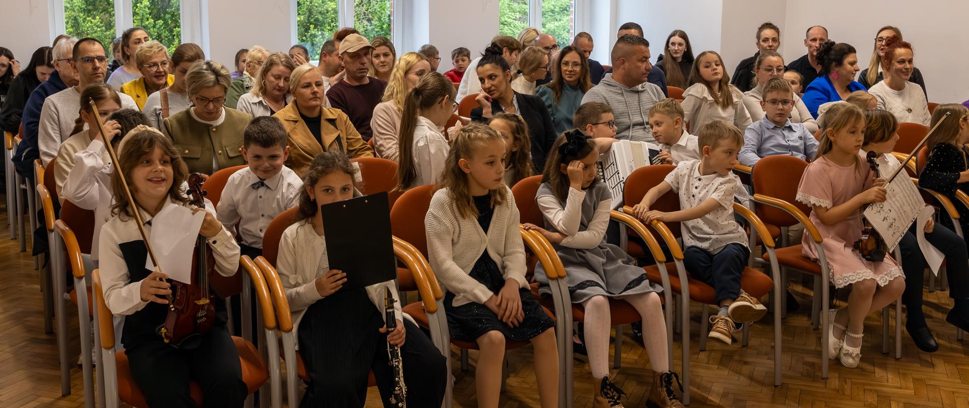 Uczniowie i rodzice zebrani na koncercie uczniów klas pierwszych jako publiczność. Pierwszy rząd zajmują uczniowie występujący na koncercie.