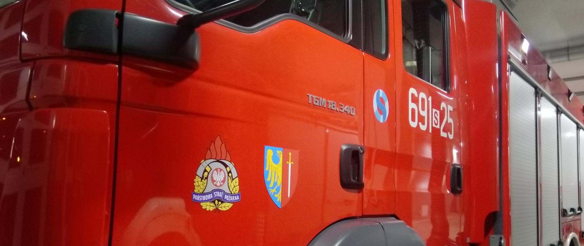 Czerwony ciężki wóz strażacki, na pierwszym planie drzwi kierowcy samochodu z logo Państwowej Straży Pożarnej oraz Miasta Żory dalej żaluzje skrytek sprzętowych samochodu