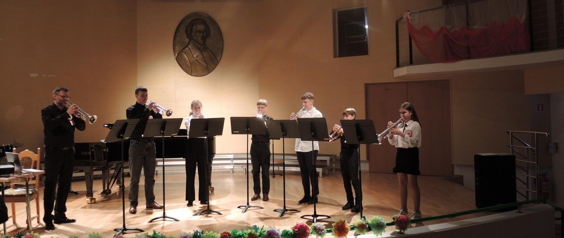Zdjęcie przedstawia zespół trębaczy stojących na scenie. Nad muzykami widnieje rzeźba Fryderyka Chopina. 