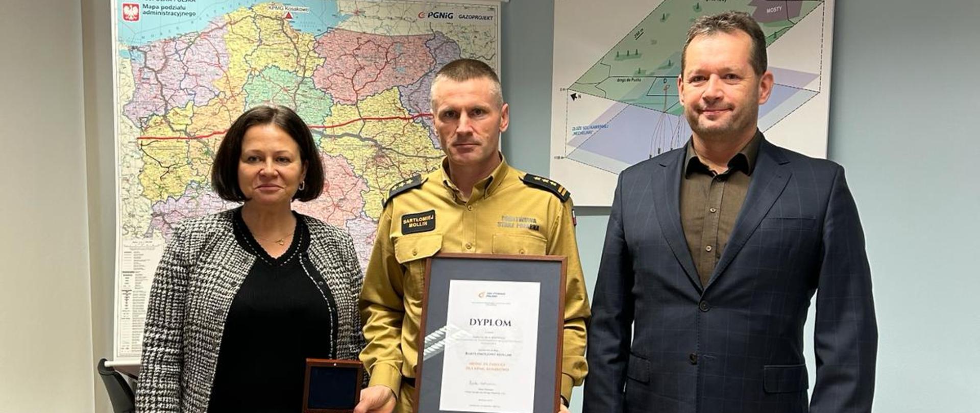 Na zdjęciu znajdują się osoby obrane w odzież cywilną oraz jedna osoba w mundurze koloru piaskowego, która trzyma dyplom oraz pamiątkowy medal.