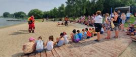Na zdjęciu strażacy oraz dzieci siedzą na plaży i opowiadają o pracy strażaka