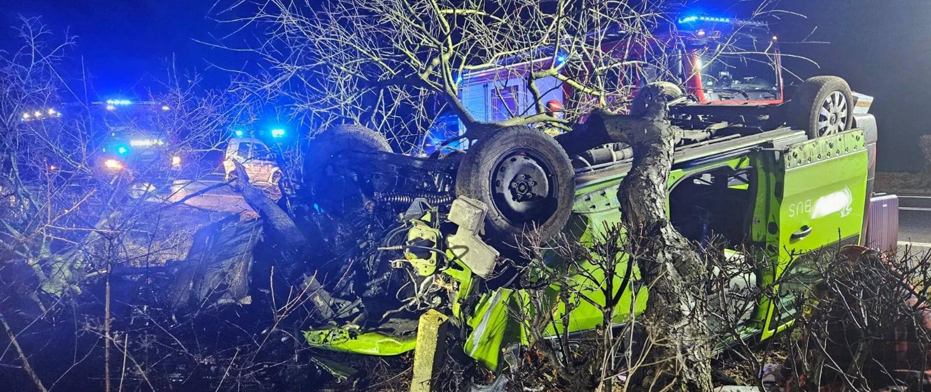 Samochód bus leżący na dachu, zniszczony przód otwarte drzwi boczne, konar drzewa na ni, w tle samochody pożarnicze