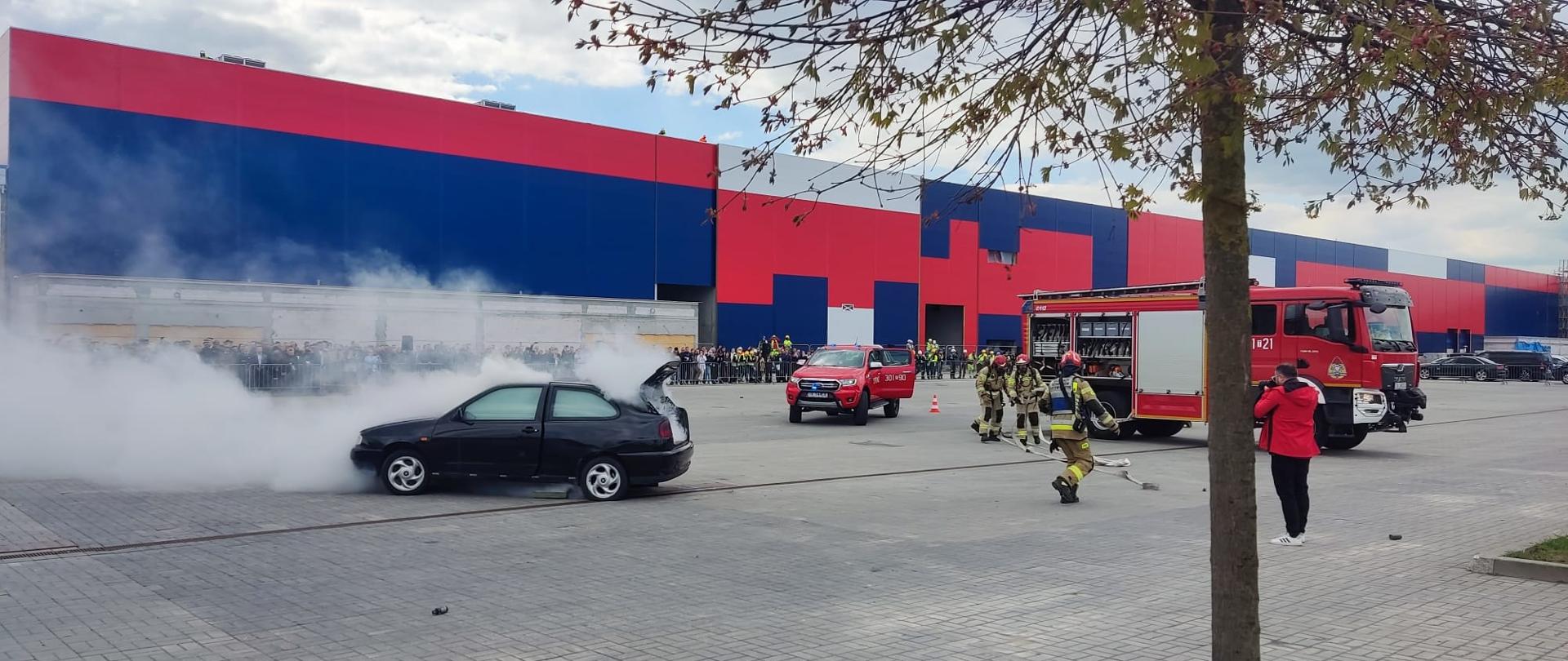 Na zdjęciu przedstawiono dynamiczny pokaz akcji gaśniczej. Płonący samochód osobowy z lewej strony w gęstym dymie, a po prawej stronie samochód gaśniczy, przy którym rozwijają się strażacy. W tle widzowie pokazu.