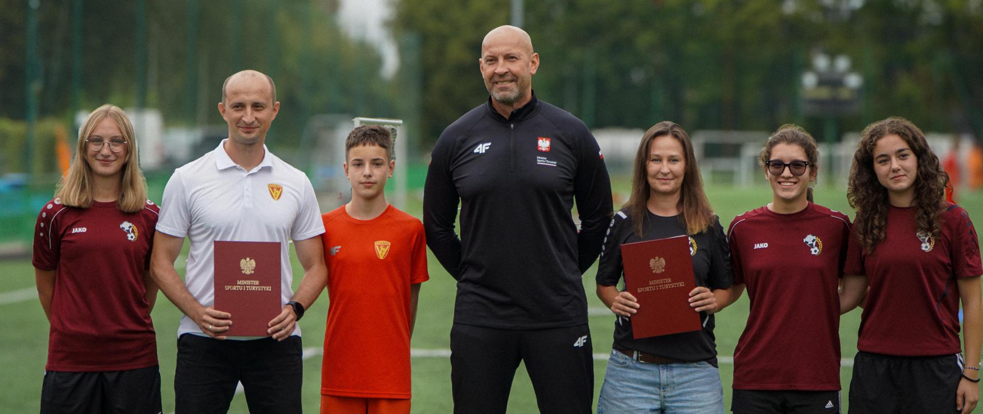 Zdjęcie. Minister sportu i turystyki Jakub Rutnicki stoi na boisku piłkarskim razem z przedstawicielami i zawodnikami klubu UKS Varsovia oraz oraz Akademii Piłkarska Diamonds Academy.