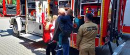 Na zdjęciu widoczny strażak w ubraniu koszarowym koloru piaskowego na tle czerwonego samochodu pożarniczego przed nim dzieci i rodzice wysłuchujący prelekcji na temat sprzętu pożarniczego i bezpieczeństwa pożarowego