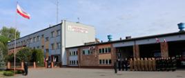Strażacy na zbiórce z okazji Dnia Flagi Rzeczypospolitej Polskiej – 2 maja. Od lewej Flaga Polski na Maszcie, poczet flagowy stoi przy maszcie i oddaje honory, po prawej strażacy w pododdziale przed garażem i Zastępca Komendanta oddają honory, wszyscy w mundurach, w tle budynek Komendy.