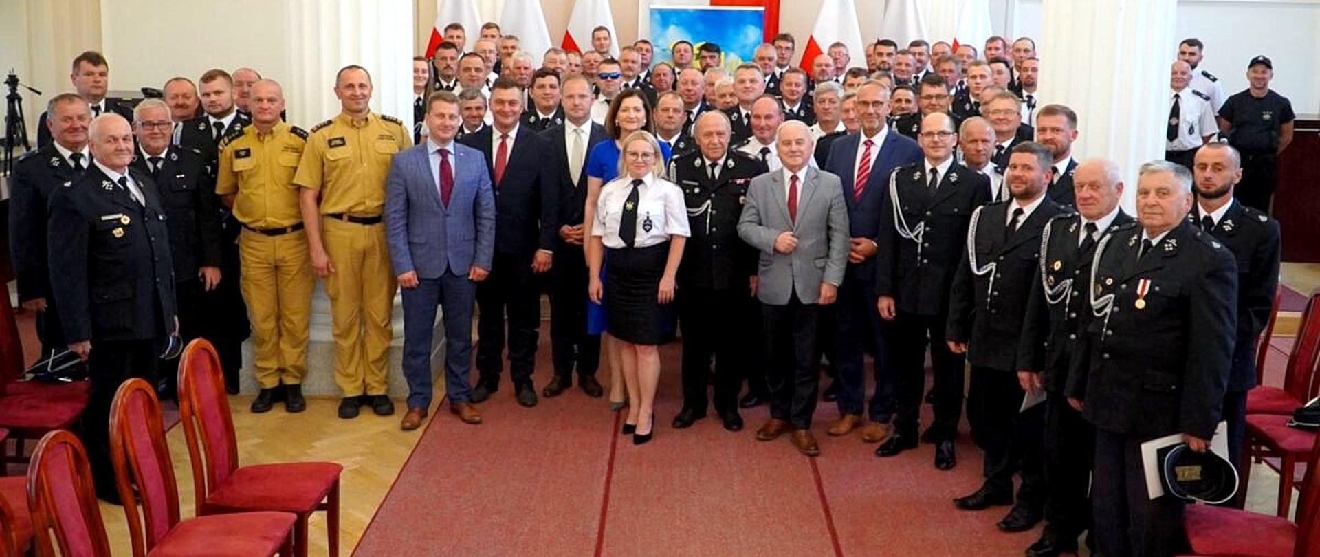 Zdjęcie zrobione wewnątrz w Sali Kolumnowej Podkarpackiego Urzędu Wojewódzkiego podczas uroczystości. Na zdjęciu stoją uczestnicy uroczystego podpisania umów, wśród nich druhowie Ochotniczej Straży Pożarnych oraz oficerowie Państwowej Straży Pożarnej. 