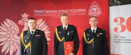 Zdjęcie zrobione w pomieszczeniu strażnicy na tle baneru Państwowej Straży Pożarnej oraz 30-lecia Państwowej Straży Pożarnej. Na pierwszym planie trzech strażaków w umundurowaniu galowym, Śląski Komendant Wojewódzki PSP nadbryg. Jacek Kleszczewski, komendant powiatowy PSP w Lublińcu oraz nowo powołany zastępca komendanta powiatowego PSP w Lublińcu.