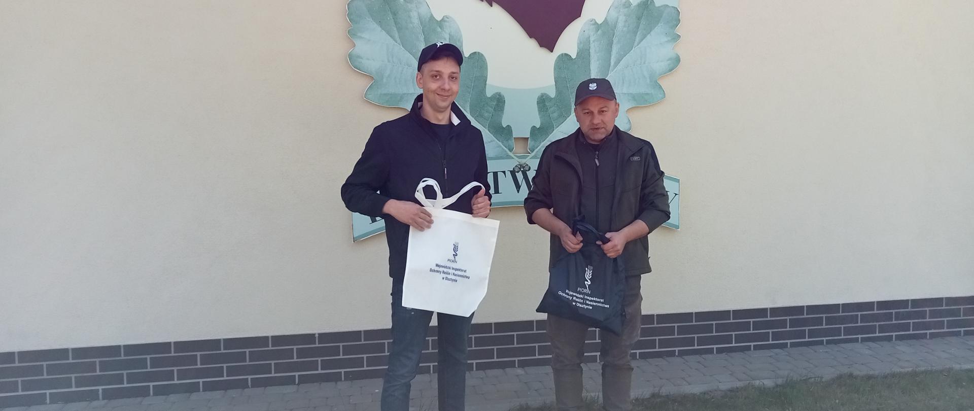 Dwóch mężczyzn stoi pozując do zdjęcia na tle logo szkółki leśnej.