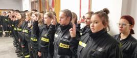 Na zdjęciu w sali konferencyjnej Komendy Powiatowej Państwowej Straży Pożarnej widoczni druhowie kończący szkolenie i składający ślubowanie ubrani w ubrania koszarowe w czarnym kolorze podczas uroczystego apelu kończącego szkolenie.