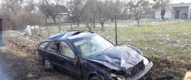 Widać uszkodzony ciemny samochód po dachowaniu, znajduje się w rowie, w tle widać drzewa w ogrodzie