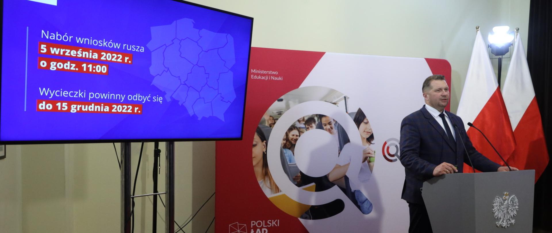 Minister Czarnek przemawia przy mikrofonie za nim ekran z prezentacją programu Poznaj Polskę 