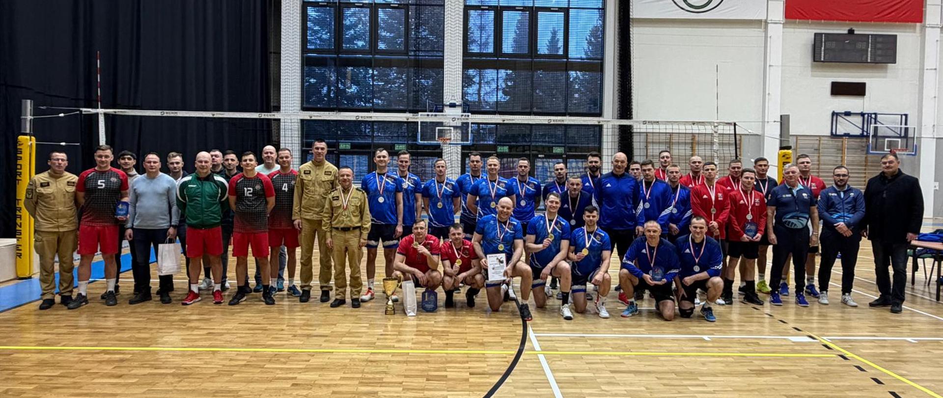 Duża grupa uczestników turnieju siatkarskiego – zawodnicy w strojach sportowych (czerwonych, niebieskich, czarnych) oraz osoby w mundurach i strojach cywilnych – pozuje wspólnie na środku hali sportowej pod siatką.
