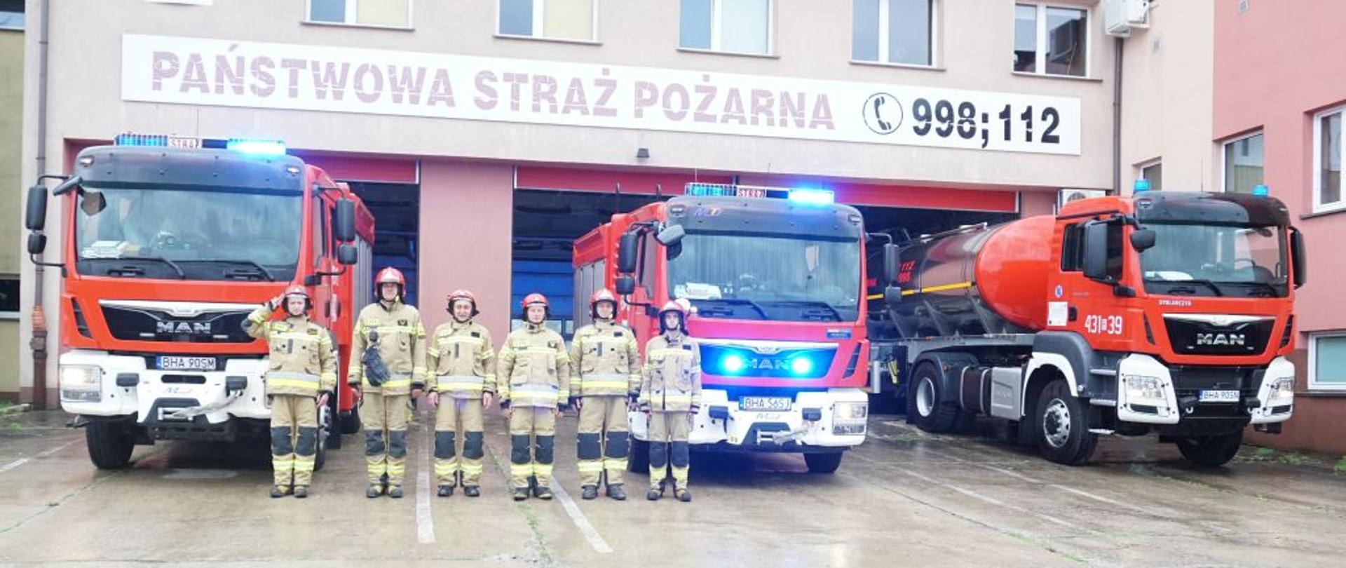 Na tle trzech czerwonych strażackich wozów bojowych stoi sześciu strażaków w żółtych mundurach bojowych. W samochodach błyskają niebieskie sygnały świetlne.