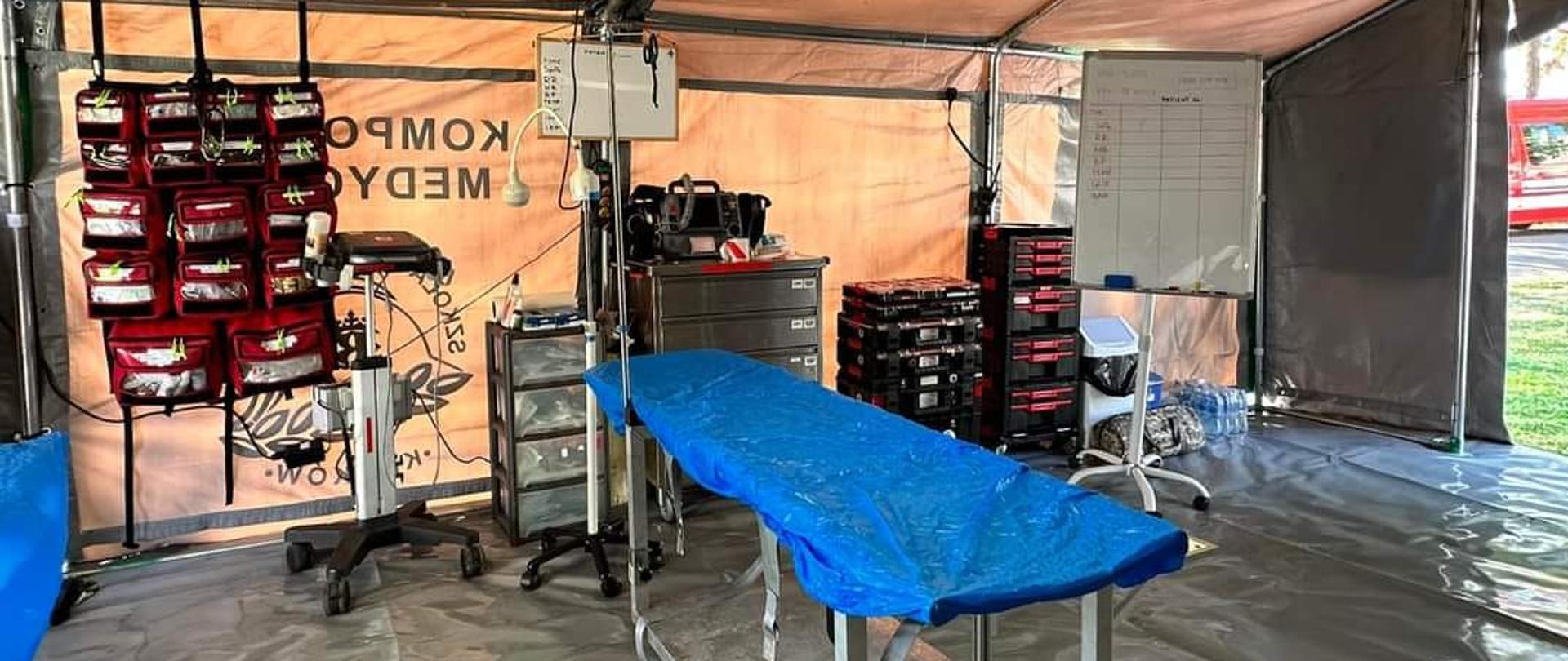 W namiocie znajduje się stół zabiegowy. Za nim wyposażenie medyczne, torby, tablica do pisania i oświetlenie.