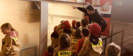 Na zdjęciu widzimy strażaka, który pokazuje grupie dzieci uczestniczącej w zajęciach, jak rozprzestrzenia się zadymienie w trakcie pożaru.