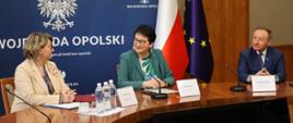 Na zdjęciu widać Wojewodę Monikę Jurek razem z Dyrektor Wydziału Polityki Społecznej Ewą Pawlinów oraz Burmistrza Olesna, Sylwestra Lewickiego. Wszyscy siedzą przy stole. Wojewoda Monika Jurek uśmiecha się i patrzy w stronę Dyrektor Ewy Pawlinów. Na stole można zobaczyć mikrofony, butelki wody oraz dokumenty. W tle widać flagę Polski oraz Unii Europejskiej a także granatowy baner z napisem "Wojewoda Opolski". 