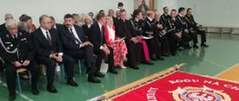 Zdjęcie przedstawia druhów oraz osoby biorące udział w obchodach 100 lecia OSP. Osoby siedzą na krzesełkach na sali gimnastycznej. Przed nimi jest stoli,, na którym leży nowy sztandar