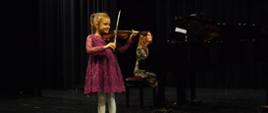 Dziewczynka gra na skrzypcach, z tyłu za nią kobieta gra na fortepianie.