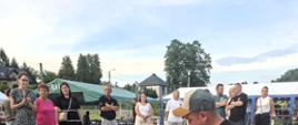 Wydarzenie plenerowe: dwóch mężczyzn klęczących na czerwonej macie na betonowym placu wykonuje resuscytację krążeniowo-oddechową na manekinie medycznym. Mężczyzna po lewej, w białej koszuli, uciska klatkę piersiową manekina. Mężczyzna po prawej, w czarnej koszulce i czapce bejsbolowej, patrzy, instruując go. W tle, na trawiastym obszarze, stoi grupa ludzi obserwujących szkolenie, a za nimi widać zielony namiot imprezowy i domy mieszkalne. Obok manekina, na macie, leży czarna torba z apteczką i biały automatyczny defibrylator zewnętrzny (AED).
