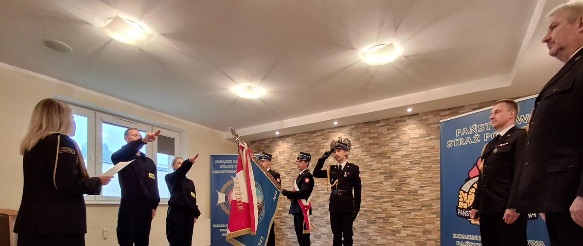 Dwóch strażaków stoi z uniesionymi prawymi rękami podczas ślubowania. Naprzeciwko nich stoi poczet sztandarowy, a z boku oficerowie PSP w mundurach galowych.