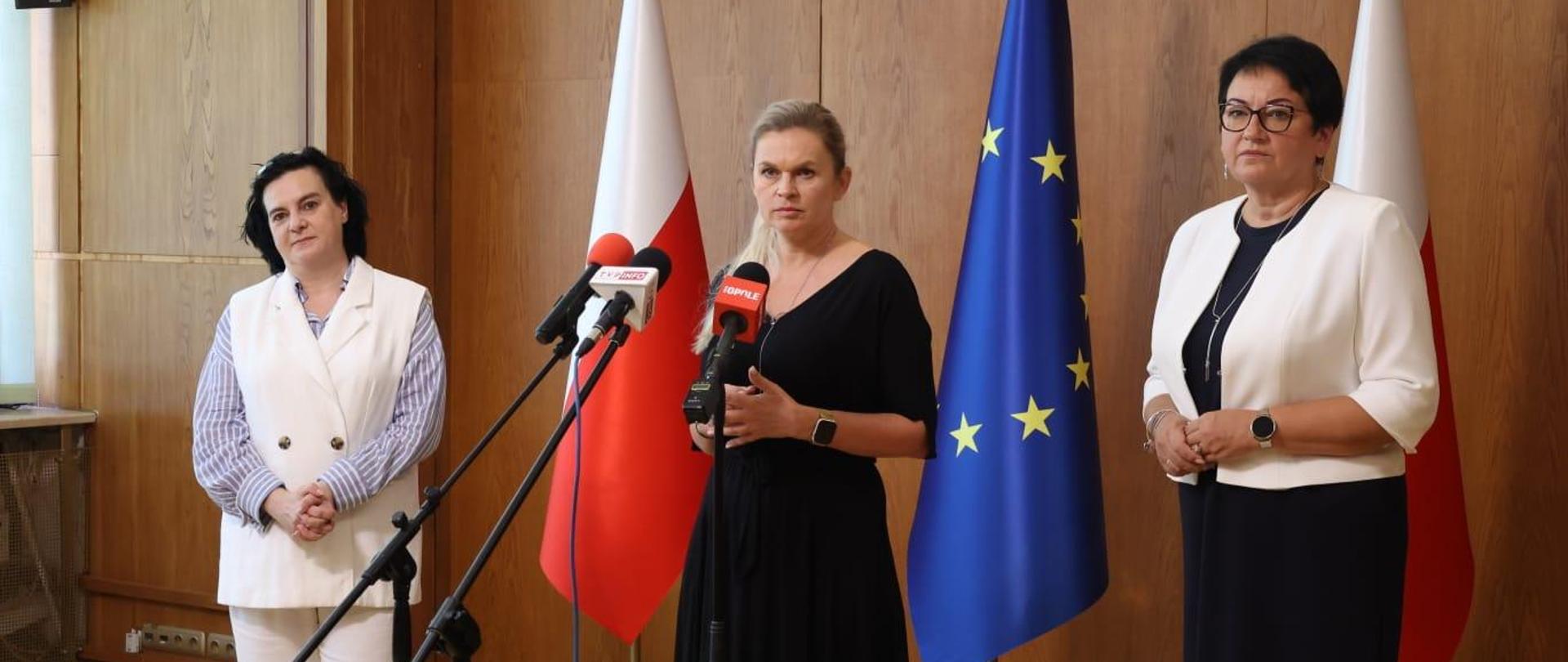 Na środku stoi ministra Barbara Nowacka. Przed nią znajdują się trzy mikrofony. Po lewej stoi pani kurator, po prawej wojewoda. Za nimi widać flagi Polski oraz Unii Europejskiej.