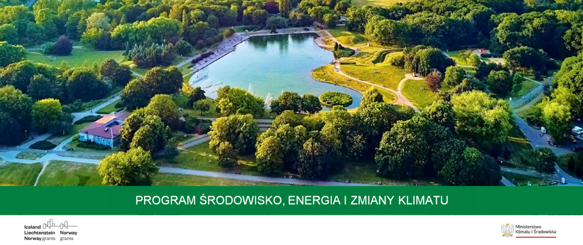 Park, na środku jezioro otoczone ścieżkami i bogatą roślinnością: drzewa, trawa. Widoczne też dwa oddalone od siebie budynki. jeden mniejszy drugi większy. 