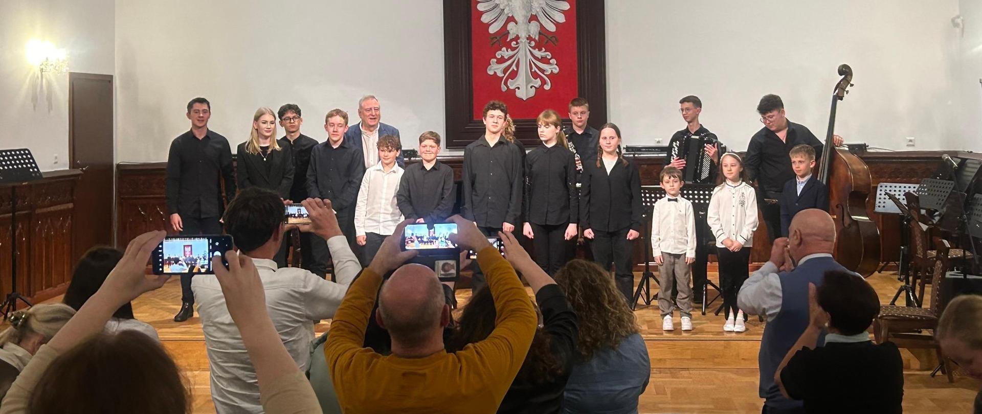 Uczniowie klasy akordeonu na scenie w sali królewskiej fotografowani licznymi telefonami uczestników koncertu