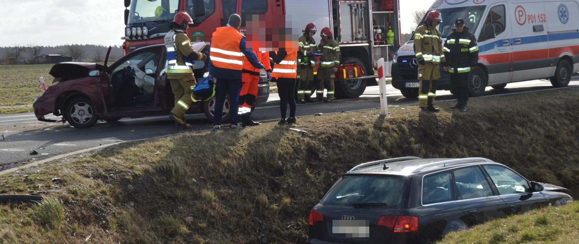 Kolizja na DW434. Skrzyżowanie dróg w okolicach Roszkówka. Na pierwszym planie audi stojące w rowie częściowo wypełnionym wodą. W tle, na jezdni drugi samochód uczestniczący w kolizji, wóz straży pożarnej oraz karetka pogotowia, strażacy i ratownicy medyczni. 