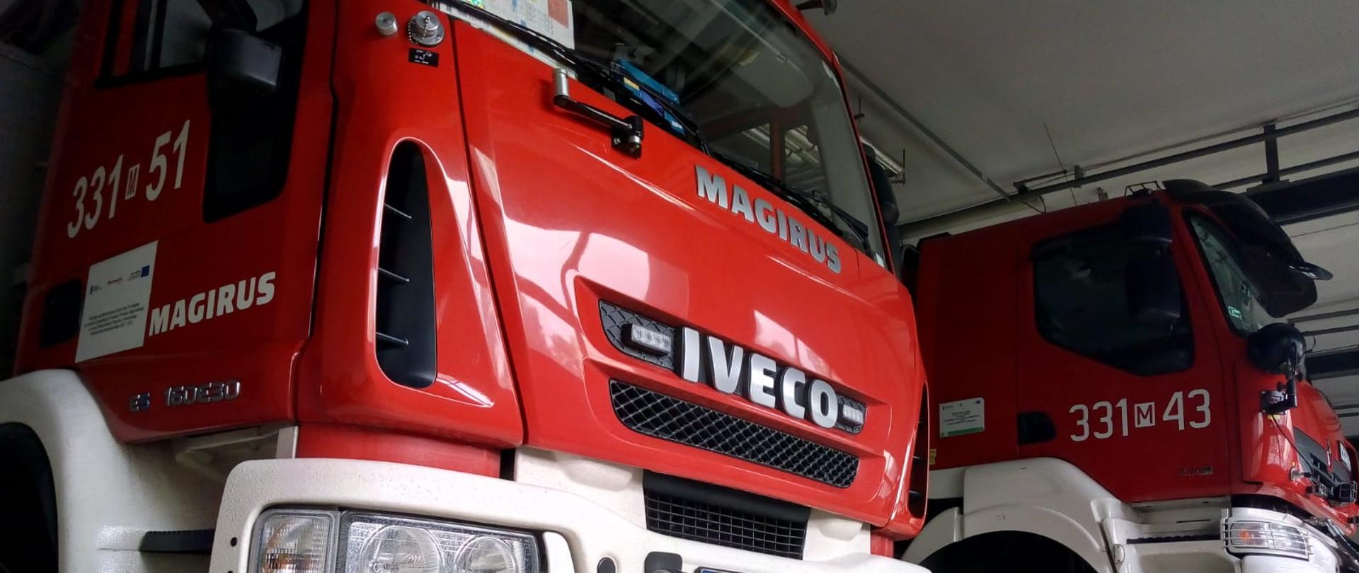 Zdjęcie wykonane w garażu jednostki Ratowniczo - Gaśniczej numer 1 w Radomiu. Na zdjęciu widać dwa pojazdy ratownicze - pojazd drabinę hydrauliczną oraz pojazd ratownictwa technicznego. Pojazdy posiadają zabudowę w kolorze czerwonym z białymi elementami podwozia. 