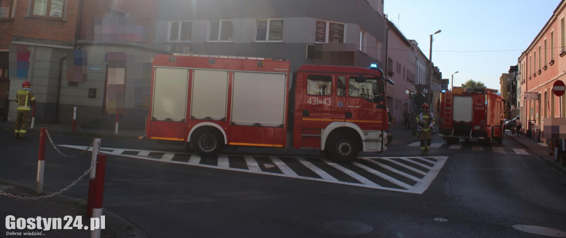 Zdjęcie przedstawia dwa samochody strażackie oraz strażaków stojących na drodze.