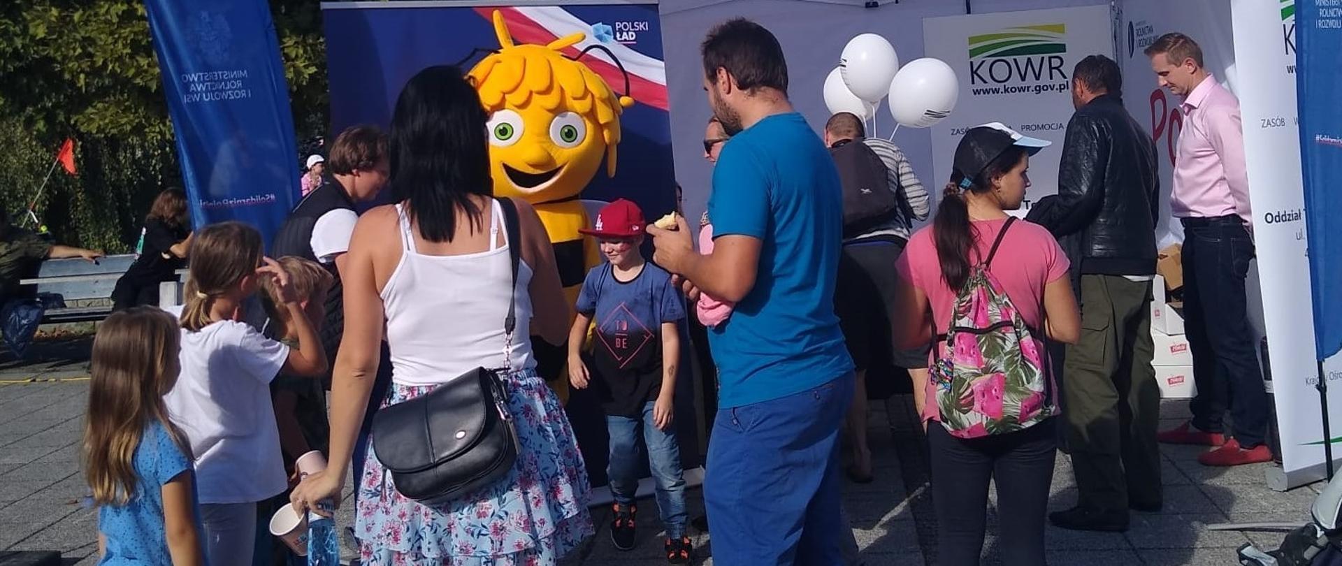 Wspólne stoisko Krajowego Ośrodka Wparcia Rolnictwa i Ministerstwa Rolnictwa i Rozwoju Wsi. W tle stoiska i plakaty promujące Polski Ład, na tle których stoi Pszczółka Maja. Przed stoiskiem rodziny z dziećmi.