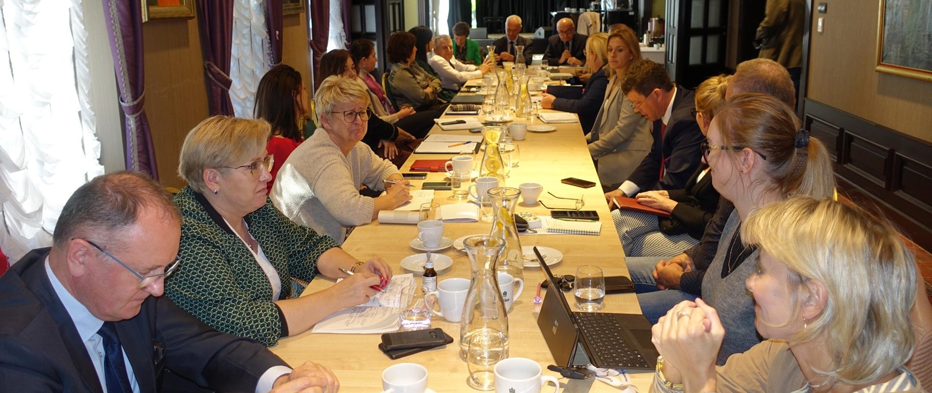 1 W centralnym miejscu zdjęcia widać długi stół, przy którym po prawej i po lewej stronie siedzi łącznie dwadzieścia osób- przeważają kobiety. Na stole znajdują się notatniki, długopisy, telefony komórkowe, laptop oraz woda w dzbankach obok której, ustawione są białe filiżanki i przezroczyste szklanki. Całość umiejscowiona jest w pomieszczeniu klimatycznie urządzonym przypominającym galerię sztuki. Na ścianie wiszą niewielkie obrazy przedstawiające postaci ludzkie. W trzech oknach po lewej stronie zawieszone są finezyjne firany z falbanami oraz purpurowe zasłony. Na tylnym planie znajdują się otwarte drewniane drzwi.