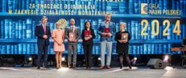 Na tle wielkiego ekranu z napisem Gala Nauki Polskiej stoi minister Wieczorek, wiceminister Mrówczyńska i cztery osoby trzymające czerwone teczki.