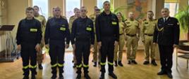 Na zdjęciu widać grupę strażaków ustawionych w dwóch rzędach w sali o drewnianej podłodze podczas ślubowania strażaków. Z przodu stoją czterej strażacy w czarnych mundurach koszarowych z naszywką „STRAŻ” na piersi, za nimi znajdują się funkcjonariusze w mundurach służbowych koloru beżowego. Po prawej stronie stoi Komendant w czarnym galowym mundurze z odznaczeniami i złotym sznurem naramiennym. W tle widoczne są duże okna, mapa Polski na ścianie oraz zielone rośliny doniczkowe.