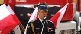 Powitanie strażaków oraz zebranych gości przez Zachodniopomorskiego Komendanta Wojewódzkiego PSP, st. bryg. Mirosława Pendera. W tle powiewają flagi Polski oraz widać samochód gaśniczy.