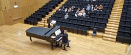 Mężczyzna gra na fortepianie, za Nim nastolatka gra na flecie, w tle widać publiczność na widowni.