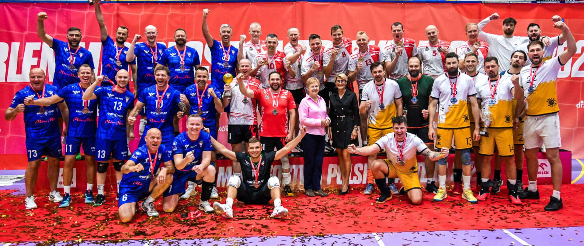 Zawodnicy drużyny Państwowa Straż Pożarna +35 pozują na podium z medalami po zdobyciu pierwszego miejsca podczas 30. ORLEN Mistrzostw Polski Masters w Piłce Siatkowej w Drzonkowie. Obok nich stoją przedstawiciele organizatorów, a w tle widoczna jest czerwona ścianka wydarzenia.