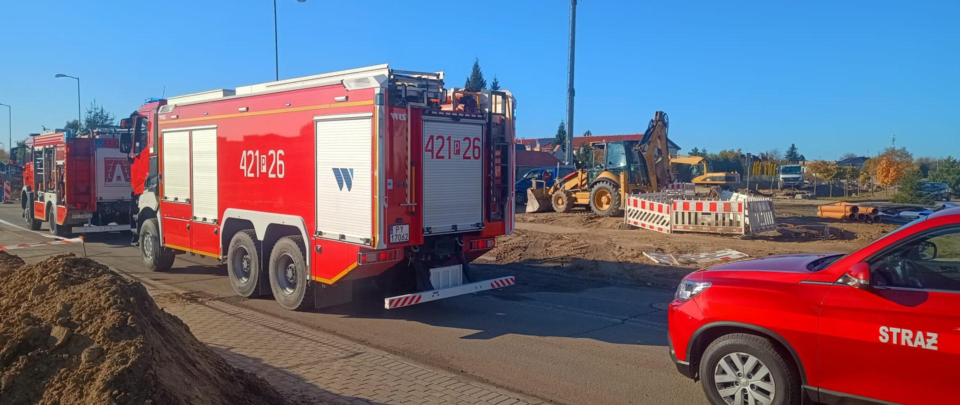 Na zdjęciu widoczne są trzy czerwone wozy straży pożarnej ustawione na drodze w pobliżu placu budowy. W tle znajduje się koparko-ładowarka, wykopy ziemne oraz biało-czerwone bariery zabezpieczające teren prac. Jest słoneczny dzień przy bezchmurnym niebie.