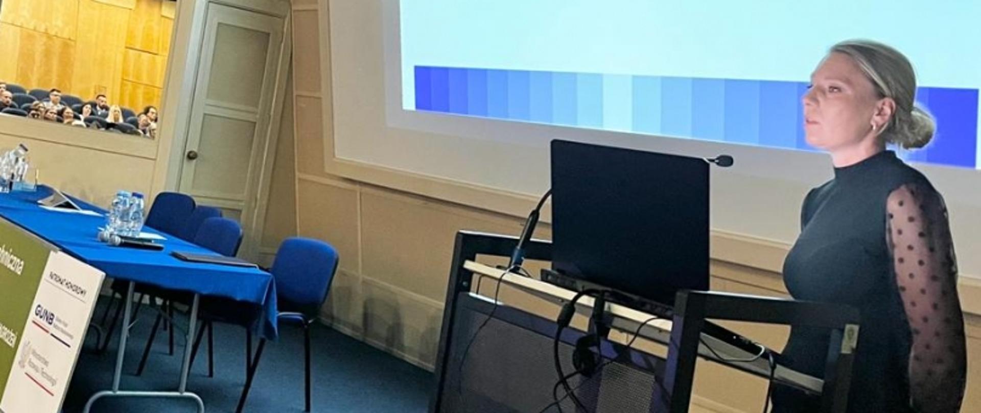 Osoba prezentująca na konferencji ECONOMIA TARS 2023, ekran z prezentacją, stół z wodą i tabliczka z nazwą wydarzenia.