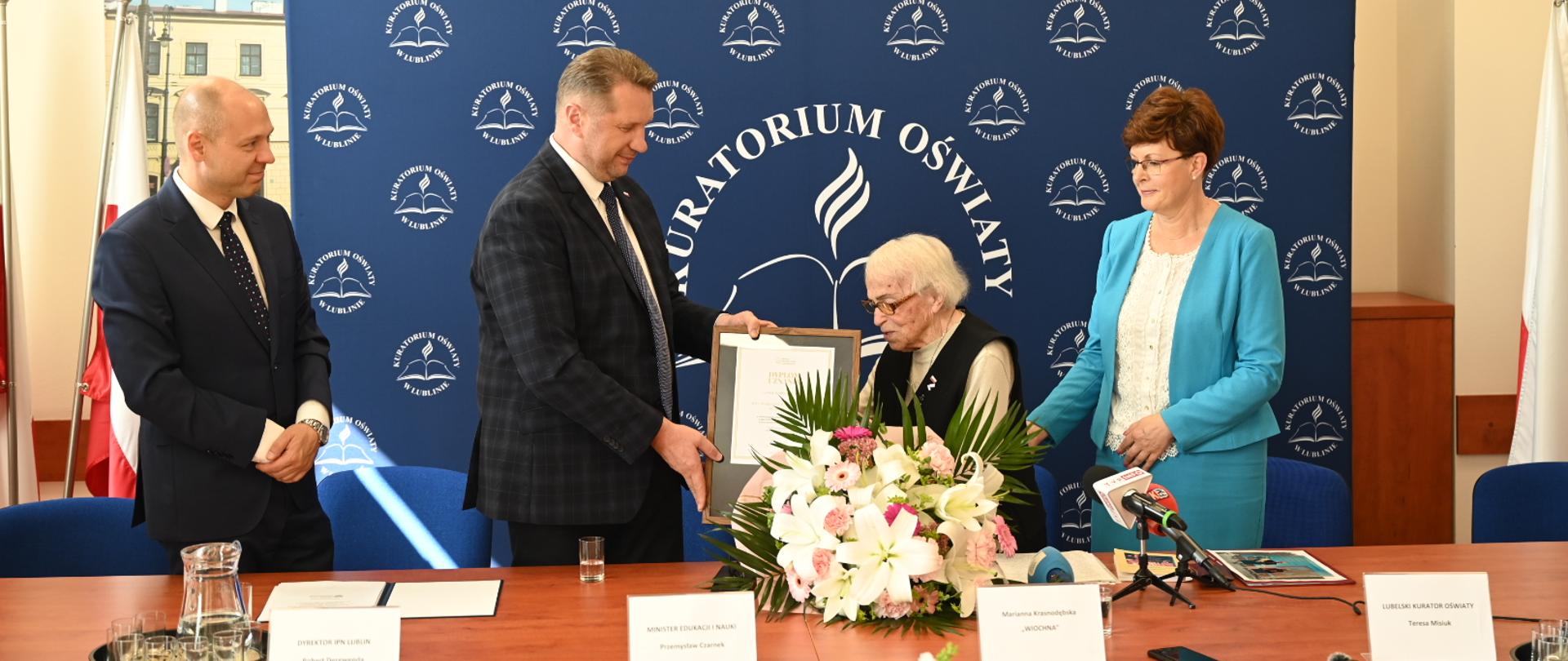 Za drewnianym stołem stoi minister Czarnek, mężczyzna w garniturze i kobieta w niebieskiej marynarce, minister podaje siedzącej przed nim kobiecie w czarnej kamizelce dokument w ramkach.