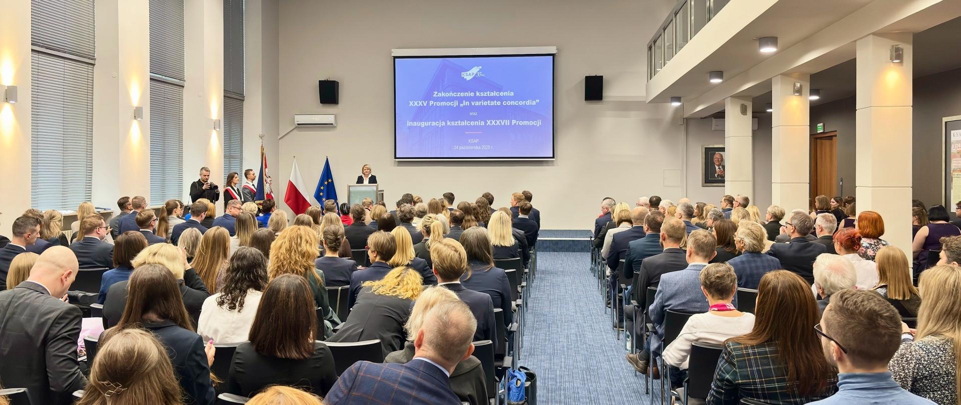 Aula wypełniona ludźmi, na mównicy dyrektorka KSAP, za nią prezentacja oraz flagi Polski i UE