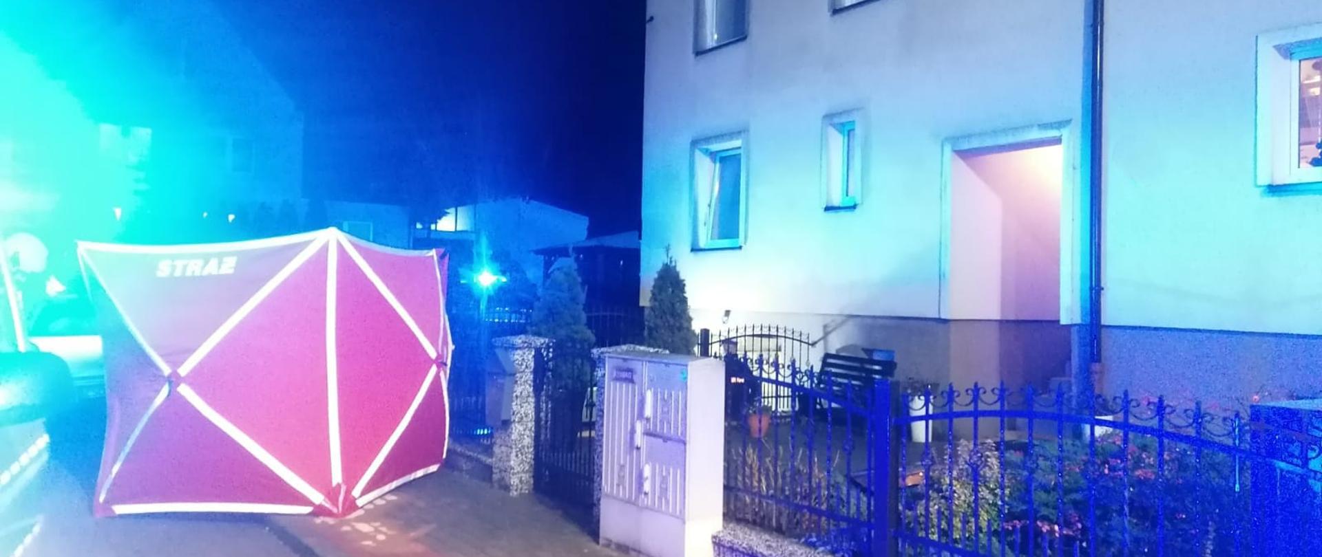 Zdjęcie przedstawia budynek wielorodzinny koloru szarego, przed nim znajduję się ogrodzenie, skrzynka elektryczna oraz czerwony parawan z napisem straż. W oknach budynku odbijają się niebieskie światła błyskowe służb ratowniczych.